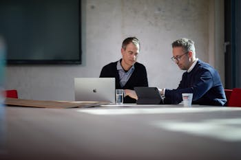 Zwei Männer sitzen nebeneinander und schauen auf Tablet und Laptop